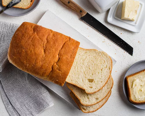 Une miche partiellement tranchée sur une planche à découper avec un couteau à pain et une tranche beurrée sur une assiette