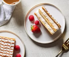  Deux morceaux de gâteau Mille-Feuille sur des assiettes, présentés avec des baies, une tasse de café et une fourchette dorée.