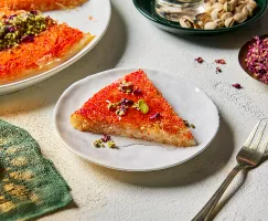 Une tranche de knafeh dans une assiette, garnie de pistaches et de pétales de fleurs comestibles, présentée avec un knafeh entier, des plats de pistaches en coque et de pétales de fleurs, et une bougie allumée.