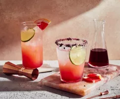 Deux verres de paloma à l'hibiscus avec un pichet de sirop simple, présentés avec un jigger et des brochettes.